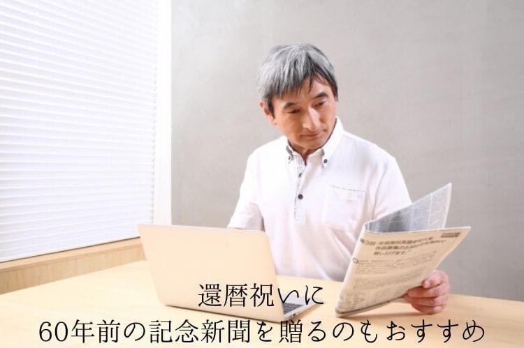 テーブルにノートパソコンを置いて手に新聞を持ち、その新聞を眺めているシニア男性