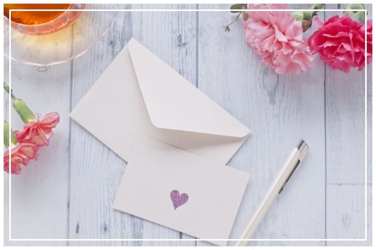 白い板と紅茶とハートが書かれたレターセット、ボールペン、カーネーションが木のテーブルに置かれている