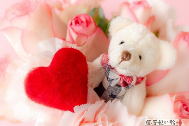 白+ピンクのカーネーションの中に白いクマのぬいぐるみと赤いハートの小物が置かれている