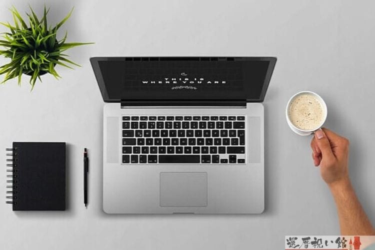 テーブルの上に置いてあるノートパソコンとノートとボールペンと観葉植物、そしてコーヒーが入ったマグカップの取っ手を握っている男性の手（腕）