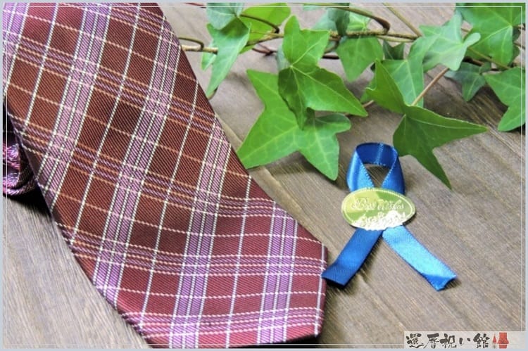 ブラウンのチェック柄のネクタイとその隣に青いリボン、植物の葉っぱが置かれている
