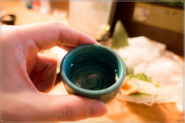 日本酒が入った緑の陶器のおちょこを手に持っている