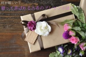 茶色の包みにこげ茶色のリボンと紫の花、メッセージカードが付いていて、その脇にピンクや紫の花が入った花束が添えられている
